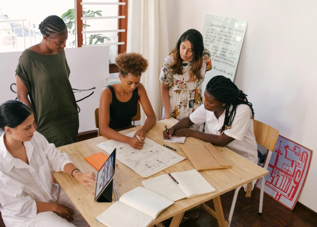 Five young black and brown women in their twenties working together, gathered around a worktable, observing and contributing to a project.