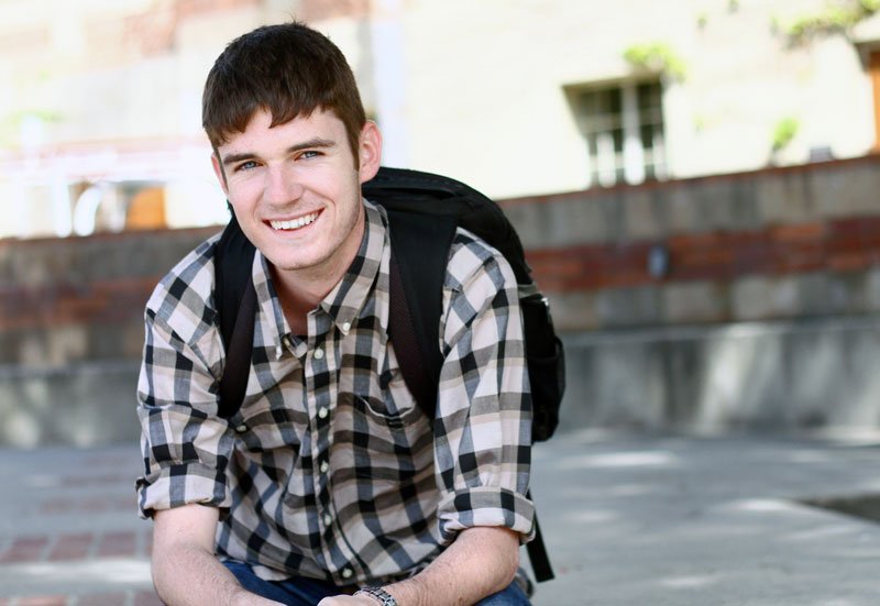 A young, white male student with short brown hair, wearing a black checkered shirt and black backpack is smiling at the camera.