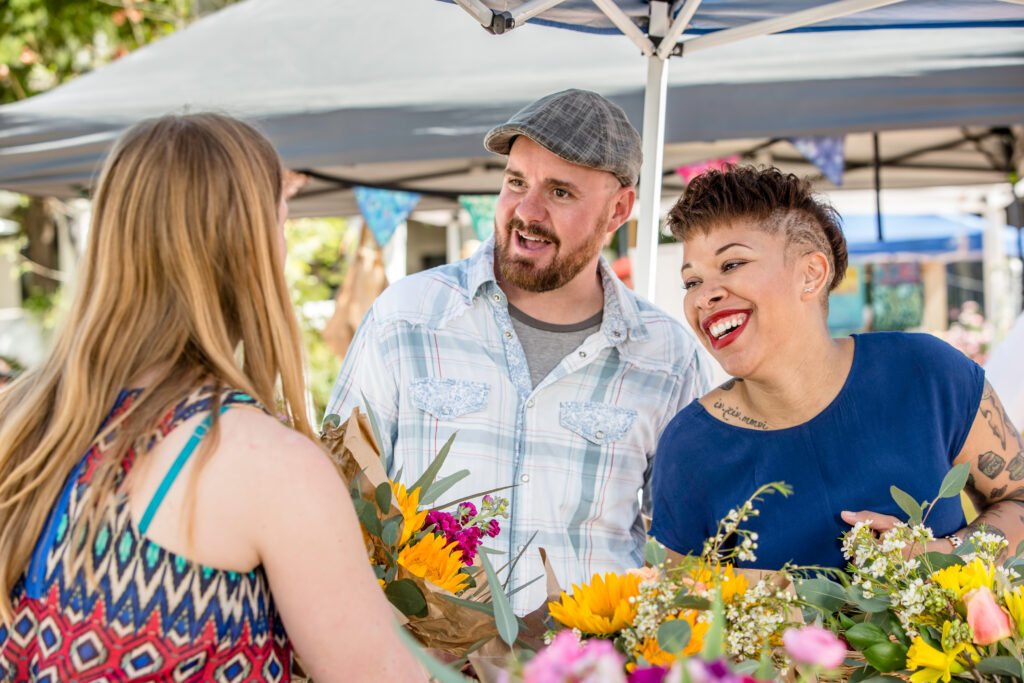 Two friendly shoppers, a bearded man and a tattooed woman chatting with a female vendor at a flower booth.